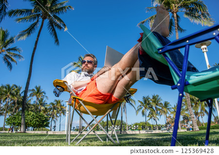 Business man using laptop at palm tree. Business trip. Man freelance worker online at beach. Businessman on summer. Online freelance work. Businessman office worker with laptop. Corporate travel plan 125369428