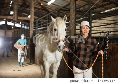 Woman horse holder leading white horse in stabling 125369447