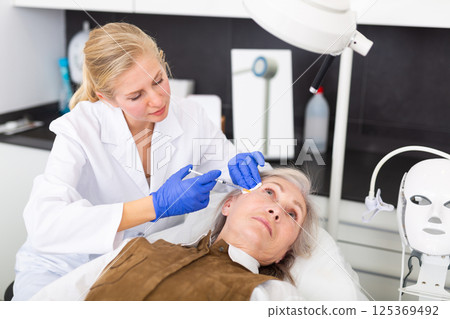 Professional beautician performing face contouring injections to aged woman Professional beautician performing face contouring injections to aged woman 125369492