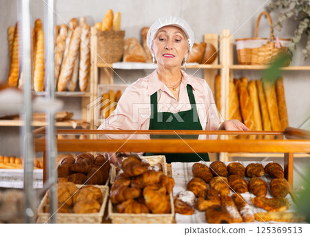 Senior woman employee puts croissants in window, arranges display of goods at bakery. Senior woman employee puts croissants in window, arranges display of goods at bakery. 125369513