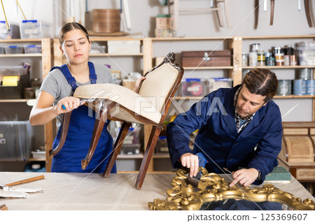 Professional male and female artisans restorers in uniform renovating antique chair mirror frame while working in repair shop Professional male and female artisans restorers in uniform renovating antique chair mirror frame while working in repair shop 125369607