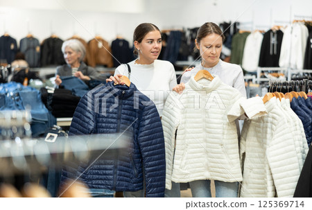 Two girlfriends choose warm fashionable jackets together in clothing store Two girlfriends choose warm fashionable jackets together in clothing store 125369714