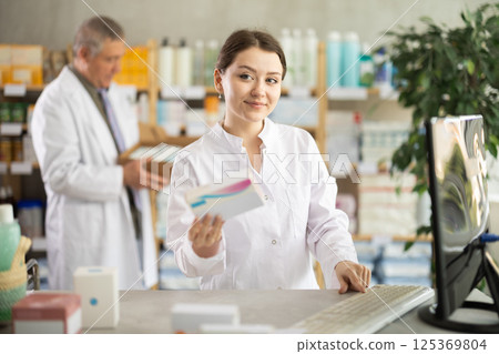 Girl pharmacist uses computer at workplace. Male colleague in background Girl pharmacist uses computer at workplace. Male colleague in background 125369804