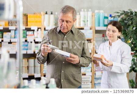 Elderly man chooses ointment in modern pharmacy. Female pharmacist helps with choosing medicine 125369870