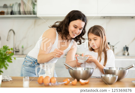 Happy mother and her daughter adding flour in metal bowl and whisking ingredients in the kitchen Happy mother and her daughter adding flour in metal bowl and whisking ingredients in the kitchen 125369876