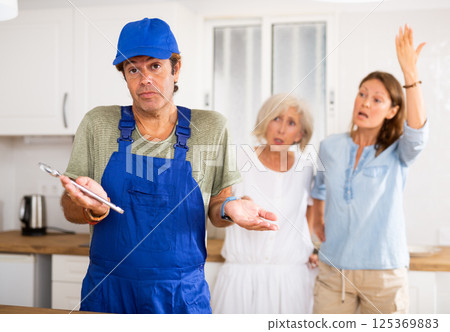 Inattentive plumber in uniform spreading his arms to the sides while being scolded by two women in the kitchen 125369883