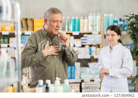 Elderly man with sore throat is looking for medicine in pharmacy. Female pharmacist is helping him 125369902