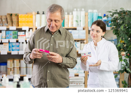 Elderly man chooses pills in modern pharmacy. Female pharmacist helps with choosing medicine 125369941