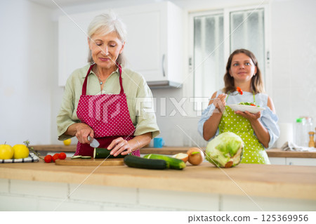 Elderly mother is preparing dinner in the kitchen. Adult daughter tries salad in background 125369956