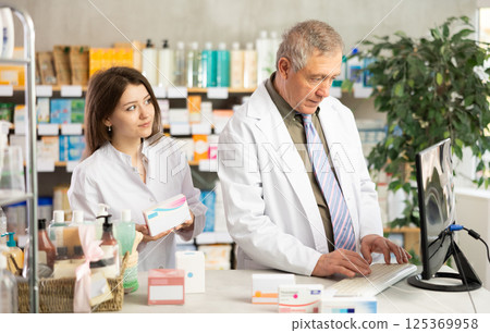 Elderly experienced pharmacist works at computer in drugstore. Young assistant helps to find pills 125369958
