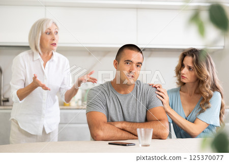 Elderly woman quarrels with couple in kitchen Elderly woman quarrels with couple in kitchen 125370077