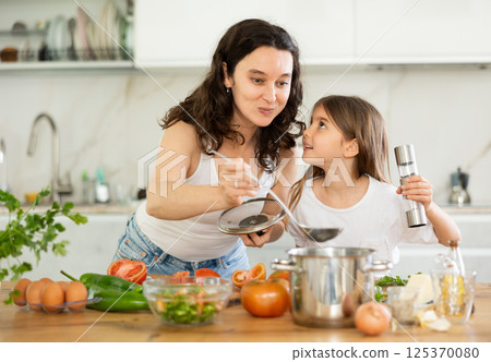 Mom and daughter taste soup in kitchen, take out portion of food from pan. 125370080