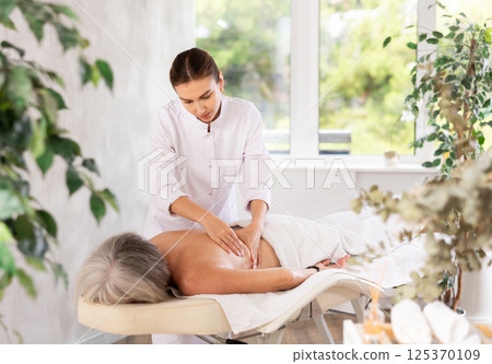 Unrecognizable elderly woman lies face down on couch and enjoy spa treatments, shoulder massage Unrecognizable elderly woman lies face down on couch and enjoy spa treatments, shoulder massage 125370109
