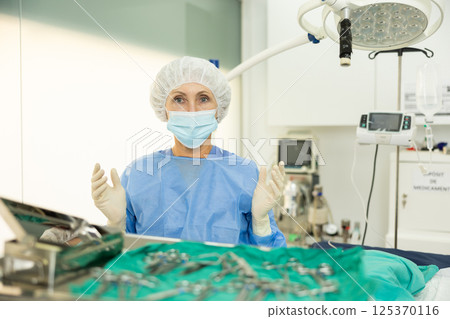 Mature woman doctor in entire perfect surgical suit stands with raised hands in gloves Mature woman doctor in entire perfect surgical suit stands with raised hands in gloves 125370116