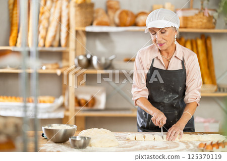 Senior woman works in bakery as baker, cuts dough into portions, form pieces of dough to create buns 125370119