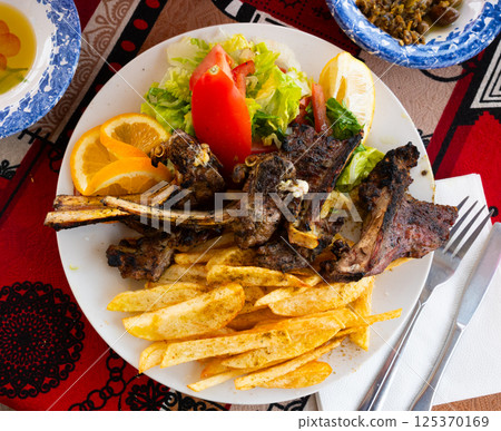 Delicious browned grilled lamb ribs served with crispy french fries and lettuce with tomato on plate, decorated with lemon. Delicious browned grilled lamb ribs served with crispy french fries and lettuce with tomato on plate, decorated with lemon. 125370169