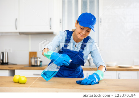 Professional female cleaner in uniform using spray chemistry on the kitchen countertop 125370194