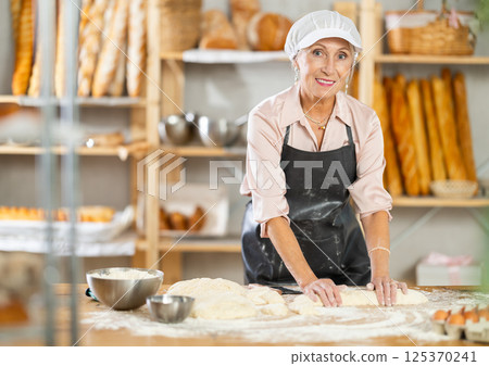 Senior woman works in bakery as baker, kneads dough, works with flour. 125370241