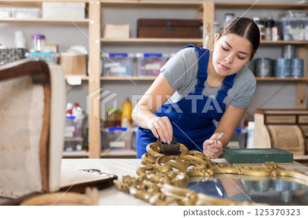 Skilled female furniture repair specialist in uniform restoring Victorian mirror frame during workday in workshop 125370323