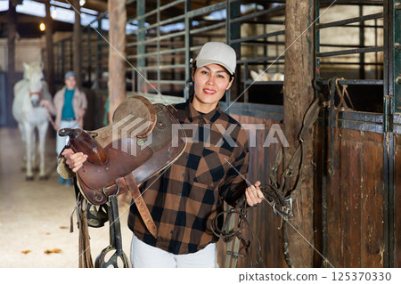 Asian female riding instructor walking in stable with saddle in hands 125370330