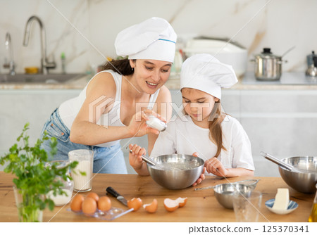 Mom and girl prepare dough for baking, mom adds salt to bowl, girl mixes ingredients with whisk. 125370341