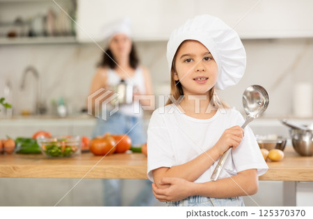 Little girl in chefs hat and white T-shirt stands in kitchen near table 125370370