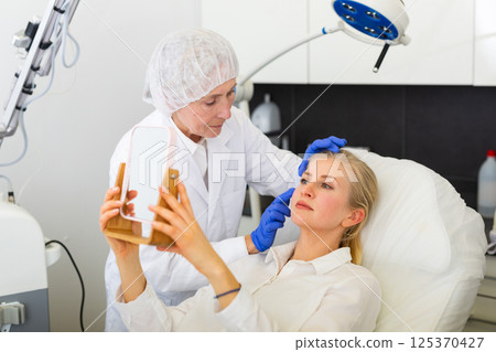 Woman evaluating results of cosmetological procedures with doctor Woman evaluating results of cosmetological procedures with doctor 125370427