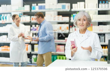 Portrait of elderly female pharmacist offering to buy medicine in pharmacy Portrait of elderly female pharmacist offering to buy medicine in pharmacy 125370506