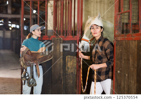 Young horsewoman holding saddle while Asian female stable keeper leading horse out of stall 125370554
