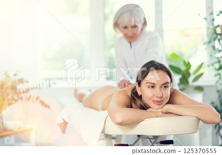 Vague mature woman actively rubs buttocks of young woman during general massage procedure. Vague mature woman actively rubs buttocks of young woman during general massage procedure. 125370555