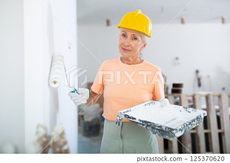Portrait of an elderly woman painter in protective helmet who paints the walls with paint roller 125370620