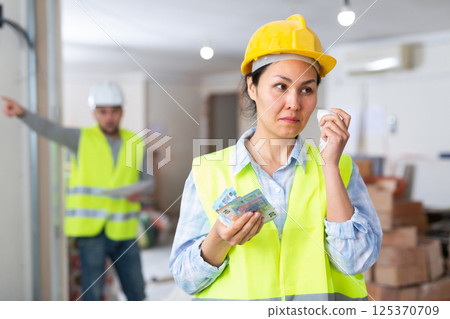 Crying construction workwoman standing at building site with banknotes Crying construction workwoman standing at building site with banknotes 125370709