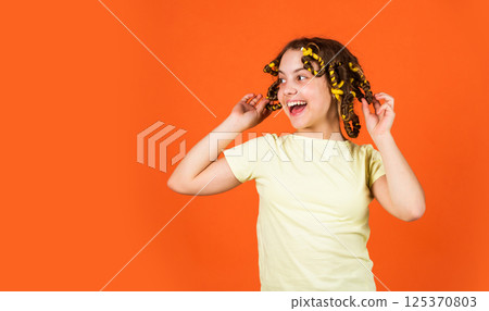 Styling tips. Small girl Curling Hair Using Curlers orange background. Adorable child hairdo. Teen hobbies. Daughter with curlers on her head laughing. Hairdresser salon. Female beauty routine 125370803