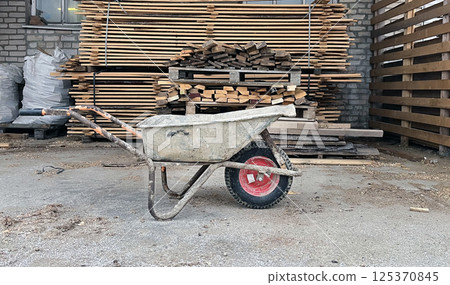 Wheelbarrow parked on a construction site with wood stacks in the background during daylight hours Wheelbarrow parked on a construction site with wood stacks in the background during daylight hours 125370845