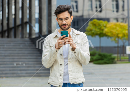 Indian man using smartphone typing text messages, reading email standing in downtown city street 125370995