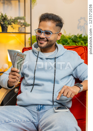 Rich happy Indian man counting money cash on smartphone calculator app, calculate income earnings 125371036