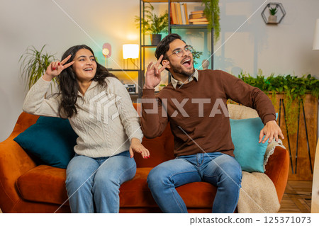 Smiling happy cheerful excited young Indian couple dancing enjoying weekend together on home couch Smiling happy cheerful excited young Indian couple dancing enjoying weekend together on home couch 125371073