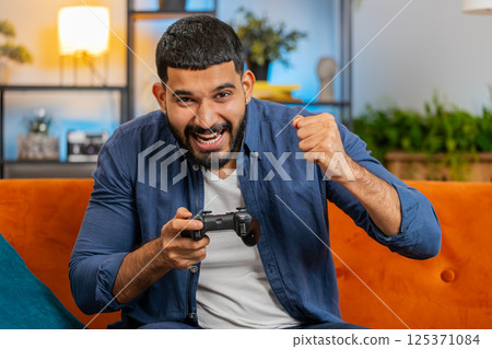 Indian man using joystick controller playing video console winning online fun game sits on home sofa 125371084