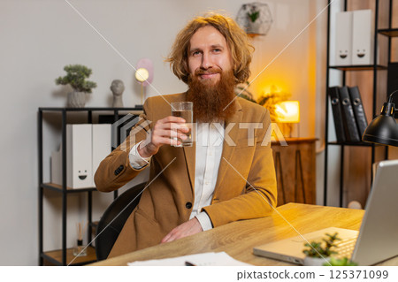 Business man sitting at workplace desk drinking water while working with laptop in home office Business man sitting at workplace desk drinking water while working with laptop in home office 125371099