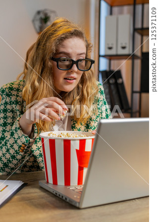 Businesswoman taking break from work wearing 3D glasses eating popcorn and watching movie at office Businesswoman taking break from work wearing 3D glasses eating popcorn and watching movie at office 125371109