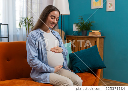 Dreamy smiling happy young pregnant woman sitting on sofa at home stroking belly expecting baby 125371184