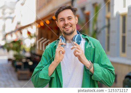 Portrait of smiling young male tourist in casual clothes with wireless headphones around neck Portrait of smiling young male tourist in casual clothes with wireless headphones around neck 125371217