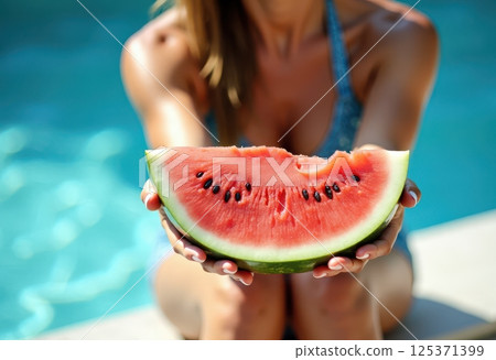 This image captures a summer scene featuring a person holding a juicy slice of watermelon by a pool. The focus is on the vibrant red watermelon with visible black seeds, held in both hands.  125371399