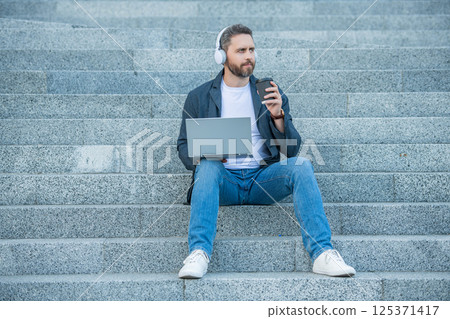 full length of man webinar online outside. photo of man webinar online on laptop with headphones full length of man webinar online outside. photo of man webinar online on laptop with headphones 125371417