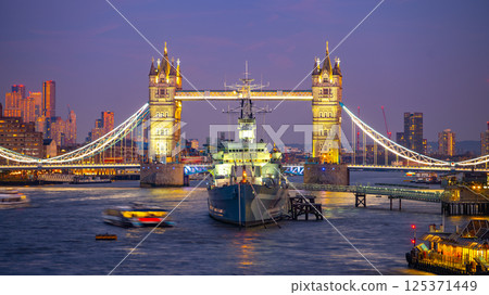 Tower Bridge stands majestically illuminated in the evening sky, while HMS Belfast rests in the Thames River. Small boats navigate the water, intensifying the vibrant scene in London. 125371449
