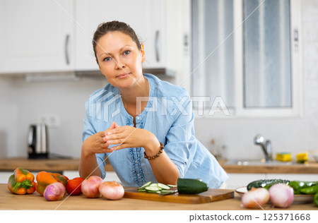 Elegant female in a blue blouse standing behind tabletop with fresh vegetables at the kitchen Elegant female in a blue blouse standing behind tabletop with fresh vegetables at the kitchen 125371668