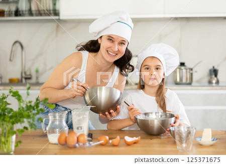 Mom and girl in white chief hats are talking in kitchen and whisking ingredients for pastry in bowl Mom and girl in white chief hats are talking in kitchen and whisking ingredients for pastry in bowl 125371675