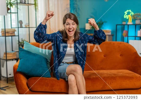 Excited happy young woman watching TV celebrating success game win dancing sitting on sofa at home 125372093