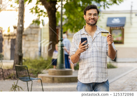 Indian young man using credit card smartphone while transferring money, purchases online outdoors Indian young man using credit card smartphone while transferring money, purchases online outdoors 125372098