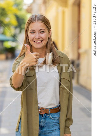 Happy young Caucasian woman looking approvingly at camera showing thumbs up like gesture on street Happy young Caucasian woman looking approvingly at camera showing thumbs up like gesture on street 125372129